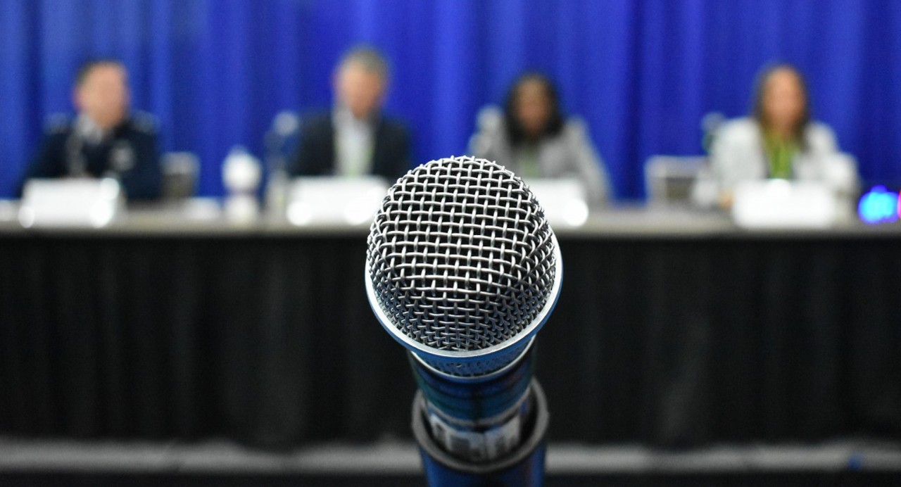Close-up of a microphone on a stand with a blurred panel of four speakers seated at a table in the background against a blue curtain. (DISA photo by David Marin)