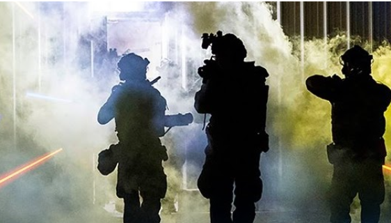Three silhouetted U.S. Army personnel move forward through dense smoke inside a building, wearing tactical gear and helmets with mounted equipment. Bright beams of colored light streak across the smoky scene, creating a high‑intensity, action-oriented atmosphere. (Defense Information Systems image by Joe Brigadier)