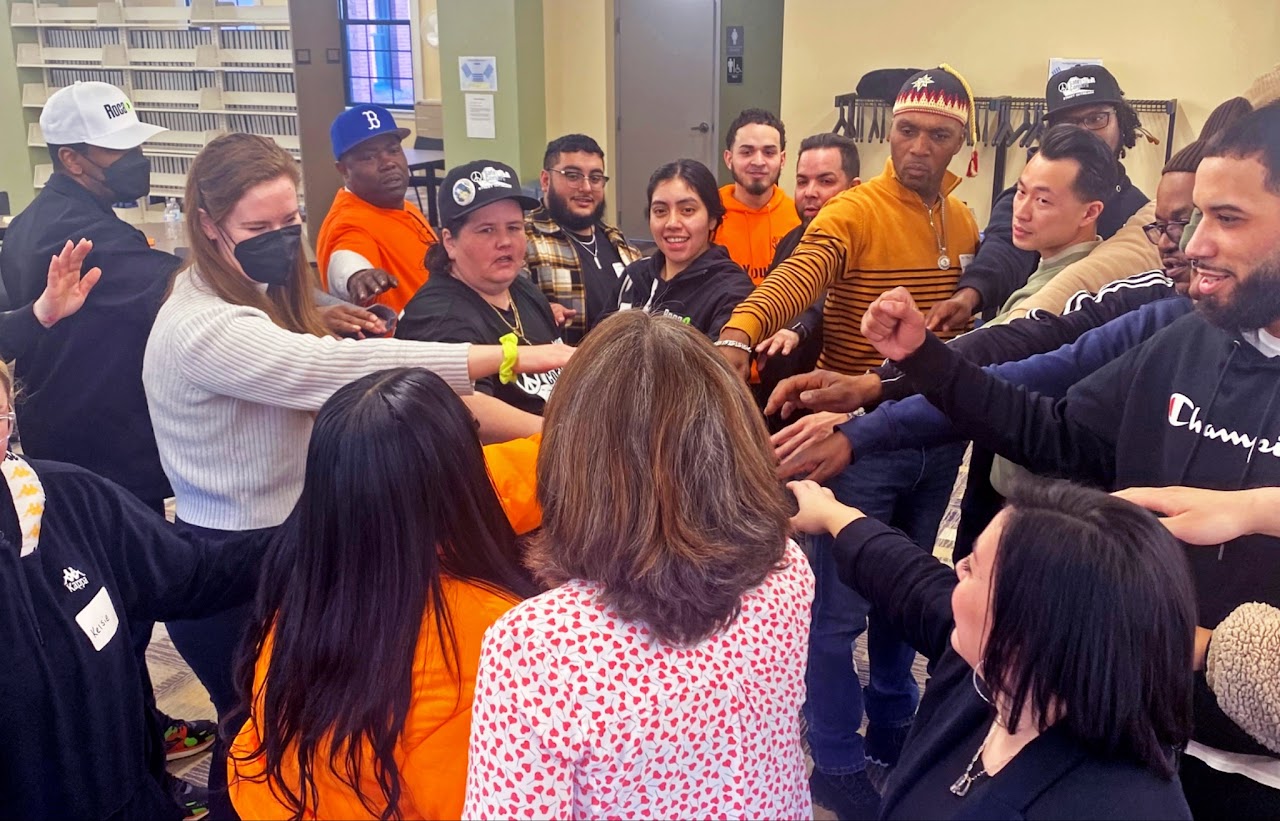 A group of people standing in a circle indoors, each extending one arm toward the center in a collective gesture. The group includes individuals wearing bright orange shirts and others in various casual clothing. Shelves and chairs are visible in the background. The setting appears to be a community space or meeting room. (Office of Justice Programs image)
