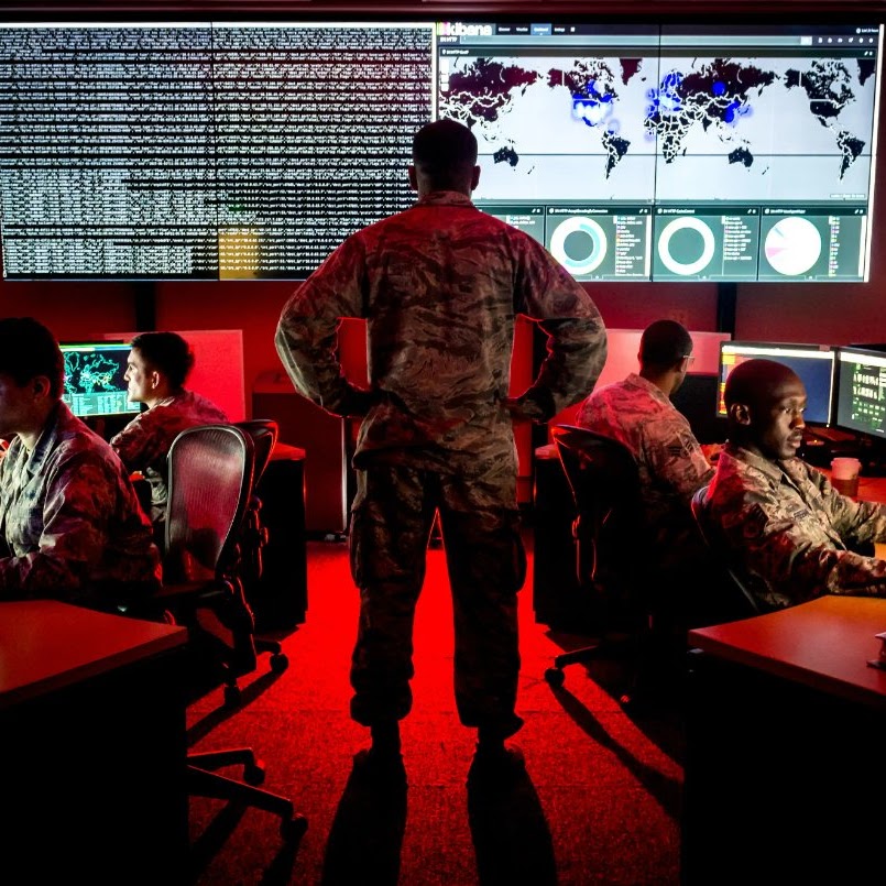 Military personnel in a cybersecurity operations center with red ambient lighting. A service member in camouflage uniform stands prominently in the center with hands on hips, facing large wall-mounted displays showing scrolling data, world maps with connection nodes, and circular analytics dashboards. Additional personnel are seated at computer workstations on either side, monitoring various screens. (Department of Defense image)