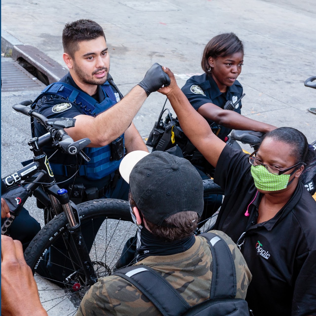 A group of people are gathered on a city sidewalk near bicycles labeled “POLICE.” Two individuals wearing dark uniforms stand beside the bicycles. Another individual wearing a face mask and a dark shirt, partially obscured, extends an arm upward in a friendly gesture, connecting hands with one of the uniformed individuals who is also reaching out. A person wearing a cap and backpack stands in the foreground with their back to the camera. (Office of Justice Programs image)