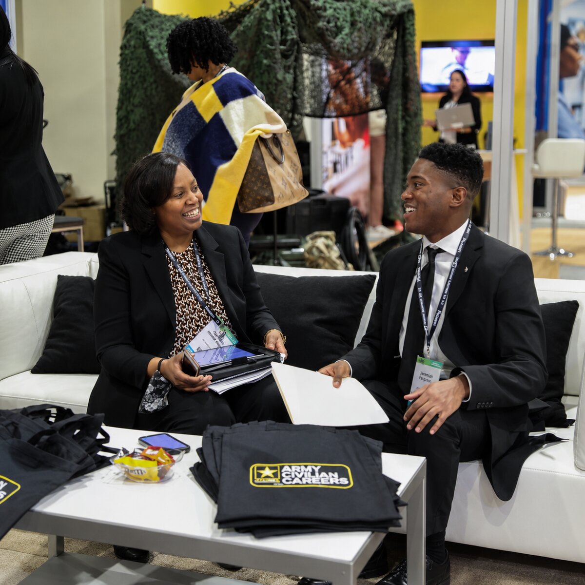 Two young professionals in business attire sit on a modern white sofa engaged in conversation at a career fair. A woman in a black blazer holds a tablet while smiling at a man in a black suit with a striped tie. On the table in front of them are promotional materials including a bag with “ARMY CIVILIAN CAREERS” branding. (U.S. Army image by Lillian Putnam)